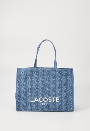 Blaue Tragetasche mit geometrischem Muster, aus Canvas-Material, mit zwei Griffen und einem großen weißen "LACOSTE 1933"-Logo auf der Vorderseite.