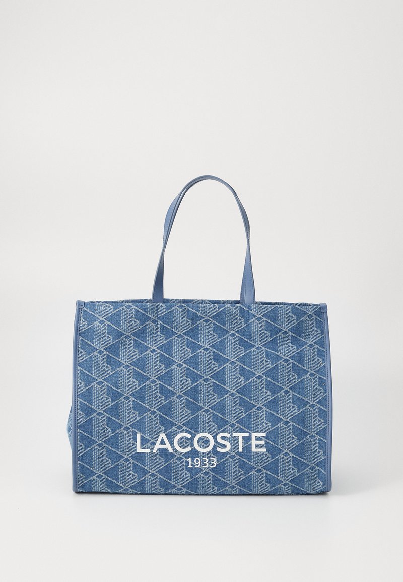 Mėlyna audinio rankinė su geometriniu raštu, turinti dvi rankenas ir didelį baltą "LACOSTE 1933" logotipą priekyje.