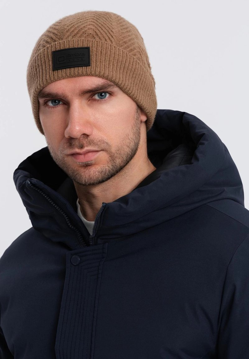 Gorro de lana marrón tejido con un patrón texturizado y un parche rectangular negro, combinado con una chaqueta azul marino con cremallera y un cuello alto.