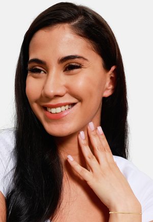 Femme avec de longs cheveux noirs portant un haut blanc, affichant des ongles manucurés de couleur rose clair. Un délicat bracelet en or orne son poignet.