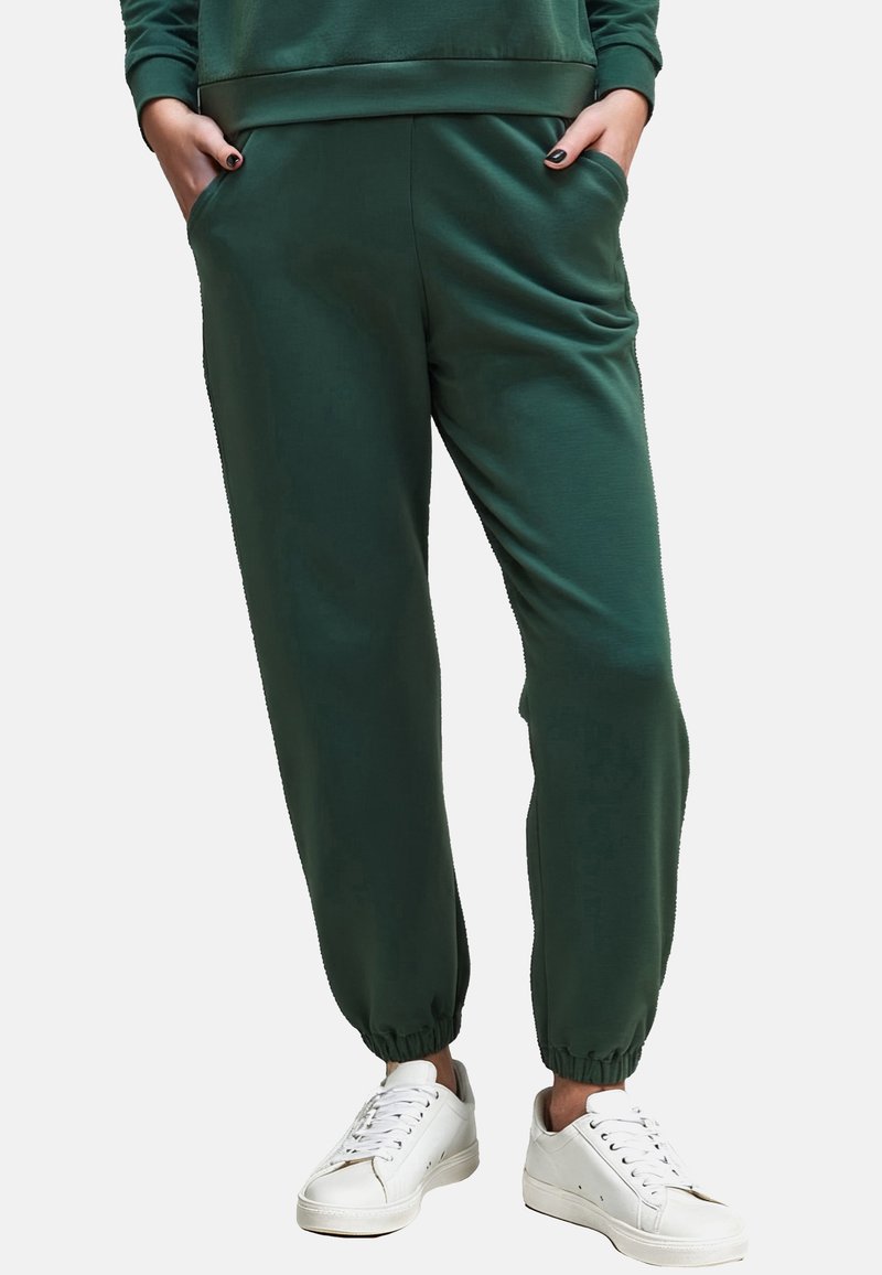 Donkergroene sweatpants met een gladde textuur, elastische tailleband en boorden. Gecombineerd met witte sneakers. Heeft zijzakken.