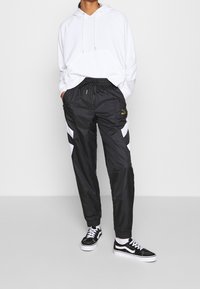 Svarta träningsbyxor med vita och svarta diagonala ränder, elastisk midja med dragkedja, parat med en vit hoodie och svarta sneakers.