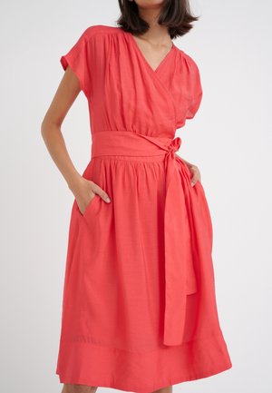 Femme portant une robe portefeuille corail avec des manches courtes, une ceinture nouée à la taille et les mains dans les poches latérales sur un fond uni.