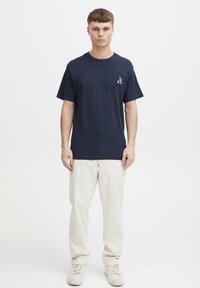 Marineblå bomull t-skjorte med rund hals og korte ermer, med et lite grafisk design på venstre bryst. Båret sammen med beige bukser.