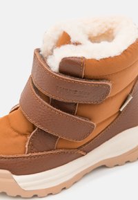 Brūni ziemas zābaki ar mīkstu baltu vilnas oderi un Velcro siksnām, ar teksturētu ādas un sintētiska materiāla augšdaļu un gumijas zoli.