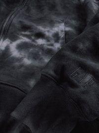 Suda preta com estampado tie-dye, com fecho e bolso frontal visível, e punhos canelados, apresentando texto bordado discreto na manga.