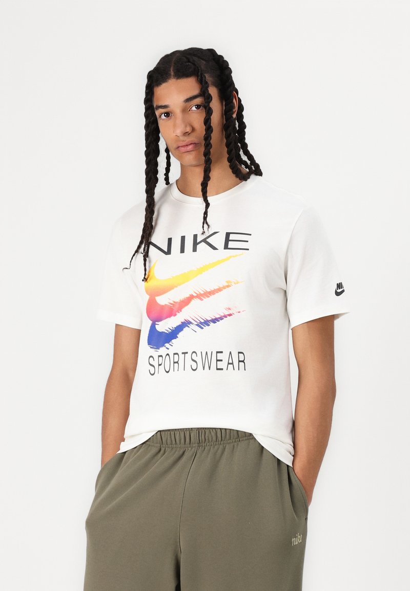 Biała bawełniana koszulka z wielokolorowym logo Nike oraz napisem "SPORTSWEAR". Krótkie rękawy i okrągły dekolt. Widać oliwkowe spodnie poniżej.