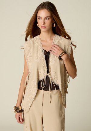 Jeune femme aux longs cheveux porte une veste en dentelle beige nouée devant, sur un top noir et un pantalon beige assorti, accessoirisée de bracelets et boucles d'oreilles en or.