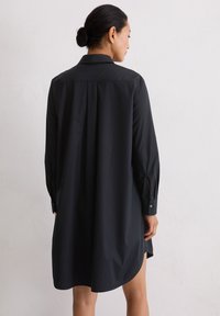 Kobieta z włosami w kokach, ubrana w luźną, długą czarną sukienkę koszulową, stoi plecami do gładkiej jasnej ściany.