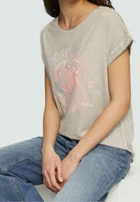 Ljusgrå bomullströja med korta ärmar, med ett grafiskt mönster i rosa och blått med cirkulära mönster. Matchad med blå jeans.