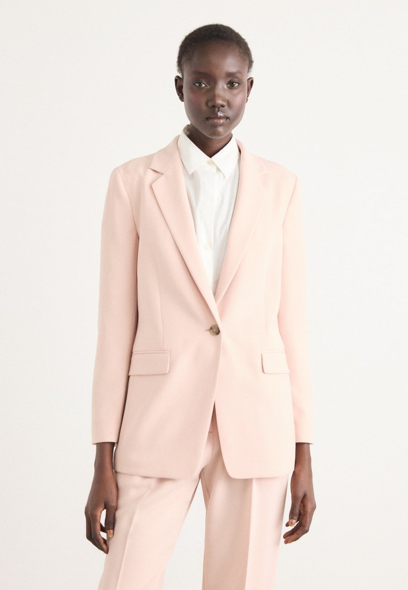 Helles rosa Blazer mit Reverskragen, einem Knopfverschluss und zwei Fronttaschen. Über einem weißen Hemd getragen. Weiche Textur und taillierte Passform.