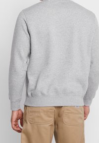 Sudadera gris con cuello redondo y puños acanalados. Presenta una textura suave y un dobladillo recto, combinada con pantalones de trabajo beige.