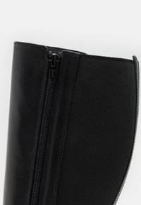 Acercamiento a un cañón de bota de cuero negro y tela con una cremallera YKK negra en el lateral, sobre un fondo blanco liso.