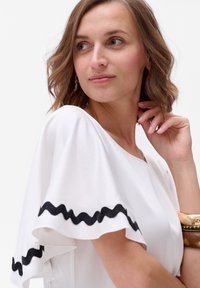 Biała bluzka z falbaniastymi rękawami, wykończona czarnym falistym wykończeniem. Modelka ma jasną karnację i brązowe włosy, trzyma złotą bransoletkę na nadgarstku.