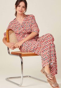 Jumpsuit de rayas rojas y crema con mangas cortas, escote en pico y piernas fluidas. Modelo sentada en una silla de madera con estructura de metal.