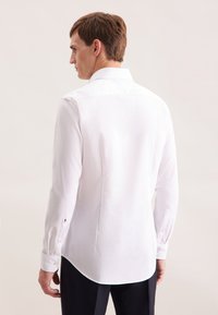 Man in een wit lange mouw overhemd en donkere broek, gezien vanuit de rug tegen een effen lichte achtergrond.