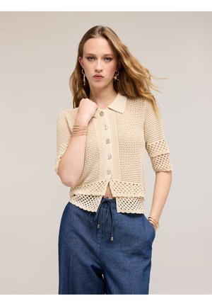 Jeune femme aux longs cheveux blonds portant un haut beige en crochet à boutons et un pantalon bleu à cordon, posant avec une main près du menton et l'autre dans la poche.