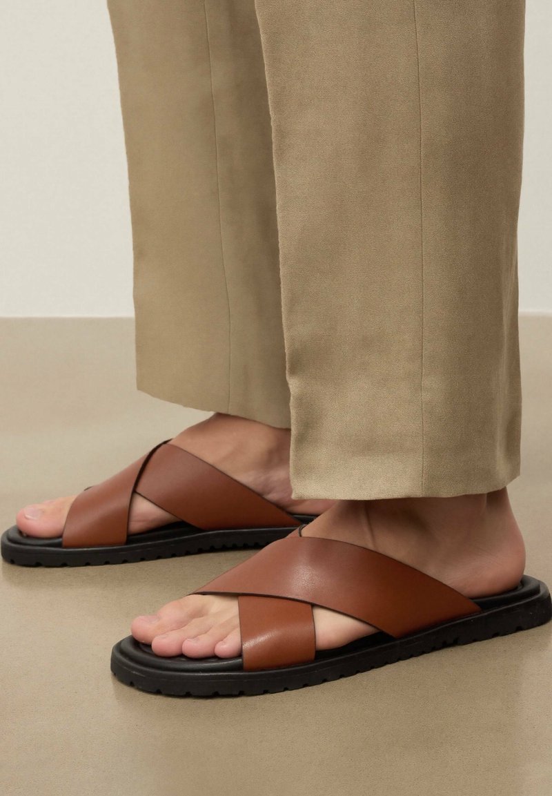Pieds chaussés de sandales en cuir marron à brides croisées avec semelles noires, associées à un pantalon beige coupé au-dessus des chevilles sur un sol neutre.