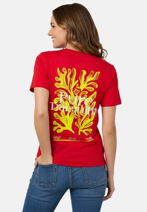 Vrouw met bruin haar, gekleed in een rood T-shirt met een gele koraalprint en de tekst "Pure Tranquility", gecombineerd met een blauwe spijkerbroek, die van opzij lacht.