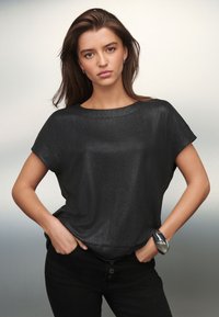 Melns īsām piedurknēm tops, izgatavots no spīdīgas auduma, ar brīvu piegriezumu un apaļu kakla izgriezumu, kopā ar melnām biksēm un sudraba rokassprādzi.