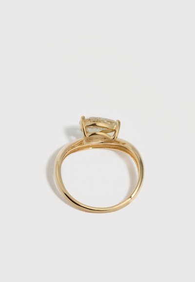 Χρυσό δαχτυλίδι με διαφανή οβάλ πολύτιμο λίθο συγκρατημένο από τέσσερις ακίδες, εμφανιζόμενο από το πλάι πάνω σε λευκό φόντο.