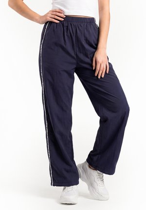 Frau trägt marineblaue, weit geschnittene Hose mit elastischem Bund und weißen Seitenstreifen, kombiniert mit weißen Sneakers und einem weißen Crop-Top.
