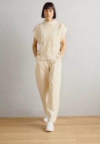 Maglione beige lavorato a maglia con dettagli a trama aperta e un design cropped, abbinato a pantaloni cream a vestibilità rilassata e sneakers bianche.