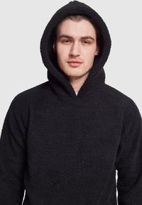 Felpa nera con cappuccio realizzata in materiale testurizzato e peloso; presenta un grande cappuccio e una vestibilità comoda, senza hardware o dettagli visibili.