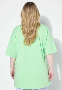 Camiseta de gran tamaño de color verde claro con mangas cortas y un dobladillo redondeado. Material de algodón, textura suave y corte holgado, vista desde atrás.