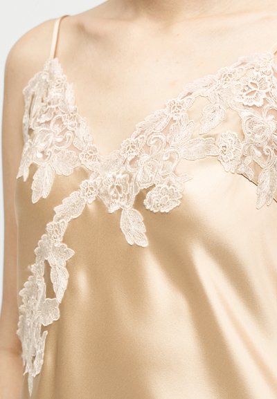 Κοντινό πλάνο χρυσής σατέν καμισόλης με περίτεχνη κρεμ δαντέλα σε σχέδια λουλουδιών κατά μήκος του ντεκολτέ και της περιοχής του στήθους.
