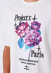 Camiseta blanca con gráficos florales vibrantes en rosa y azul con el texto: "Project x Paris" y "aventura comunitaria," impresión texturizada.