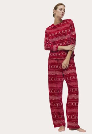 Mujer de pie descalza vistiendo una camiseta de manga larga roja y pantalones con patrones blancos de copos de nieve y geométricos.