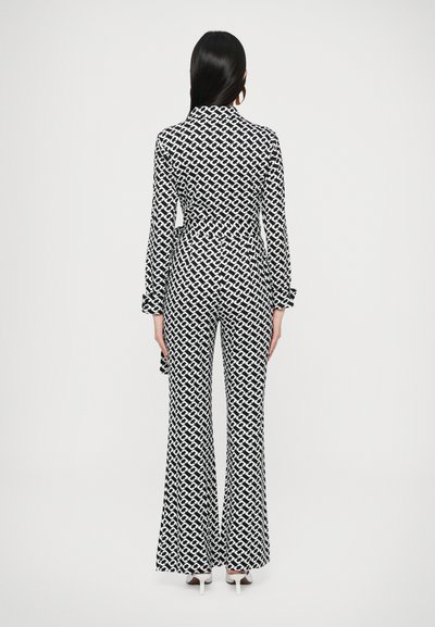 Ολόσωμη φόρμα σε ασπρόμαυρο με μοτίβα, μακριά μανίκια, ξεχειλωμένα μπατζάκια, ζώνη στη μέση και λαιμόκοψη με γιακά, που παρουσιάζει γεωμετρικό σχέδιο.