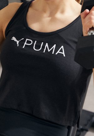 Osoba w czarnym topie Puma podnosi hantlę podczas treningu.