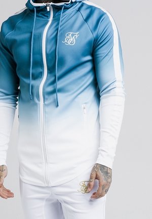 Mężczyzna w niebiesko-białej gradientowej bluzie na zamek z kapturkiem i białych spodniach, obie z ozdobnym monogramem "FS" na klatce piersiowej i udzie.