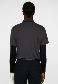 Kortärmad mörkgrå polo med en subtil textur, bärs över långsvarta ärmar, ihop med svarta byxor och en svart keps.