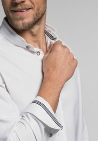 Ein weißes Hemd mit einem Kragen, der mit einem schwarzen Streifen akzentuiert ist, am Hals geknöpft und mit hochgeschobenen Ärmeln, die ein ähnliches schwarzes Detail aufweisen.