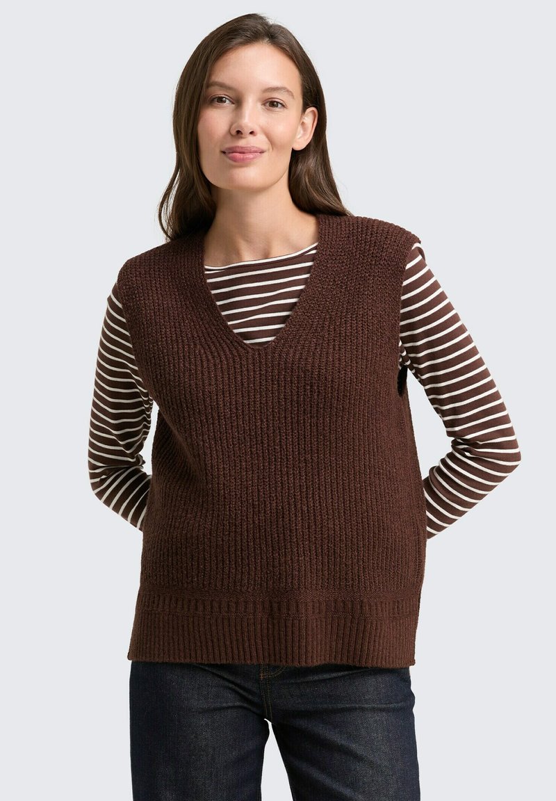 Bruine gebreide vest met een diepe V-hals, geribbelde textuur en een brede tailleband, gedragen over een bruin-wit gestreepte langemouwenshirt.