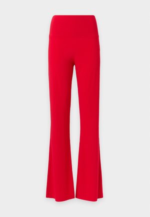 Femme portant un haut blanc Bardot à manches longues, un pantalon rouge taille haute, des sandales blanches à talons hauts, un collier en or et des boucles d’oreilles créoles.