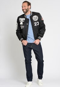 Zwarte bomberjack met badges, gestreepte manchetten en kraag, gedragen over een lichtblauwe blouse, gecombineerd met donkere jeans en witte sneakers.