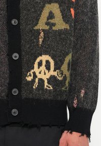 Cárdigan negro de punto con patrones gráficos multicolores, que incluyen letras y un símbolo de paz, con bordes deshilachados y cuatro botones.
