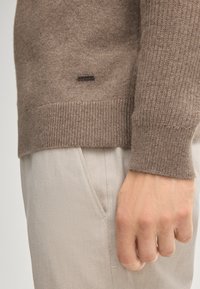 Brauner gestrickter Pullover mit gerippten Bündchen, ausgestattet mit einem kleinen schwarzen Metall-Logoschild. Kombiniert mit hellbeigen strukturierten Hosen.