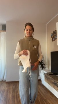 Veste en polaire avec poche zippée, de couleur beige, portée sur une chemise à manches longues de couleur claire. La personne tient un vêtement blanc avec des bretelles.