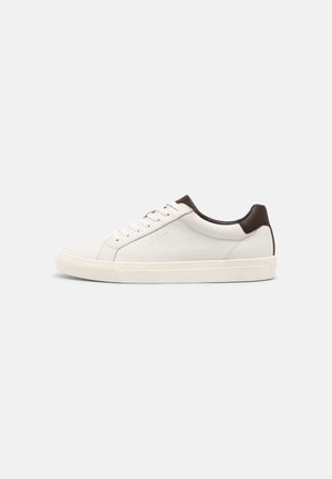 White low-top sneaker with light sole, brown heel tab, lace-up front, and subtle side logo on plain background.