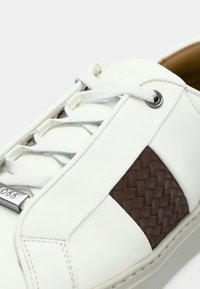 Sneaker in pelle bianca con pannello laterale in pelle intrecciata marrone, lacci bianchi e targhetta metallica sui lacci, su sfondo bianco.