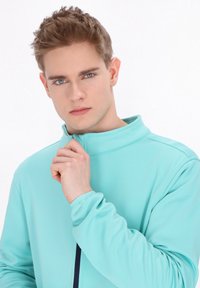 Un pullover verde menta con zip, colletto alto e maniche lunghe, realizzato in tessuto morbido e dal design aderente, indossato con le mani sul colletto.