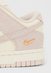 Béžový semišový a kožený podpatek tenisky se strukturovaným límcem a zlatým kovovým logem Nike ve tvaru „swoosh“ nad krémově zbarvenou podrážkou.