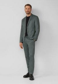 Costume texturé gris avec une coupe ajustée, associé à une chemise noire. Il présente des revers crantés et des détails subtils sur les poches, porté avec des chaussures noires.