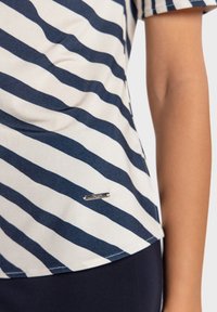 Krótka koszulka w paski granatowo-białe z metalicznym logo przy dolnym brzegu, wykonana z gładkiej tkaniny o luźnym kroju.