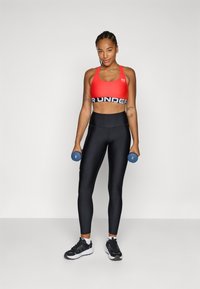 Sujetador deportivo rojo con cinturilla negra, mallas negras ajustadas y zapatillas en blanco y negro; sosteniendo mancuernas azules.